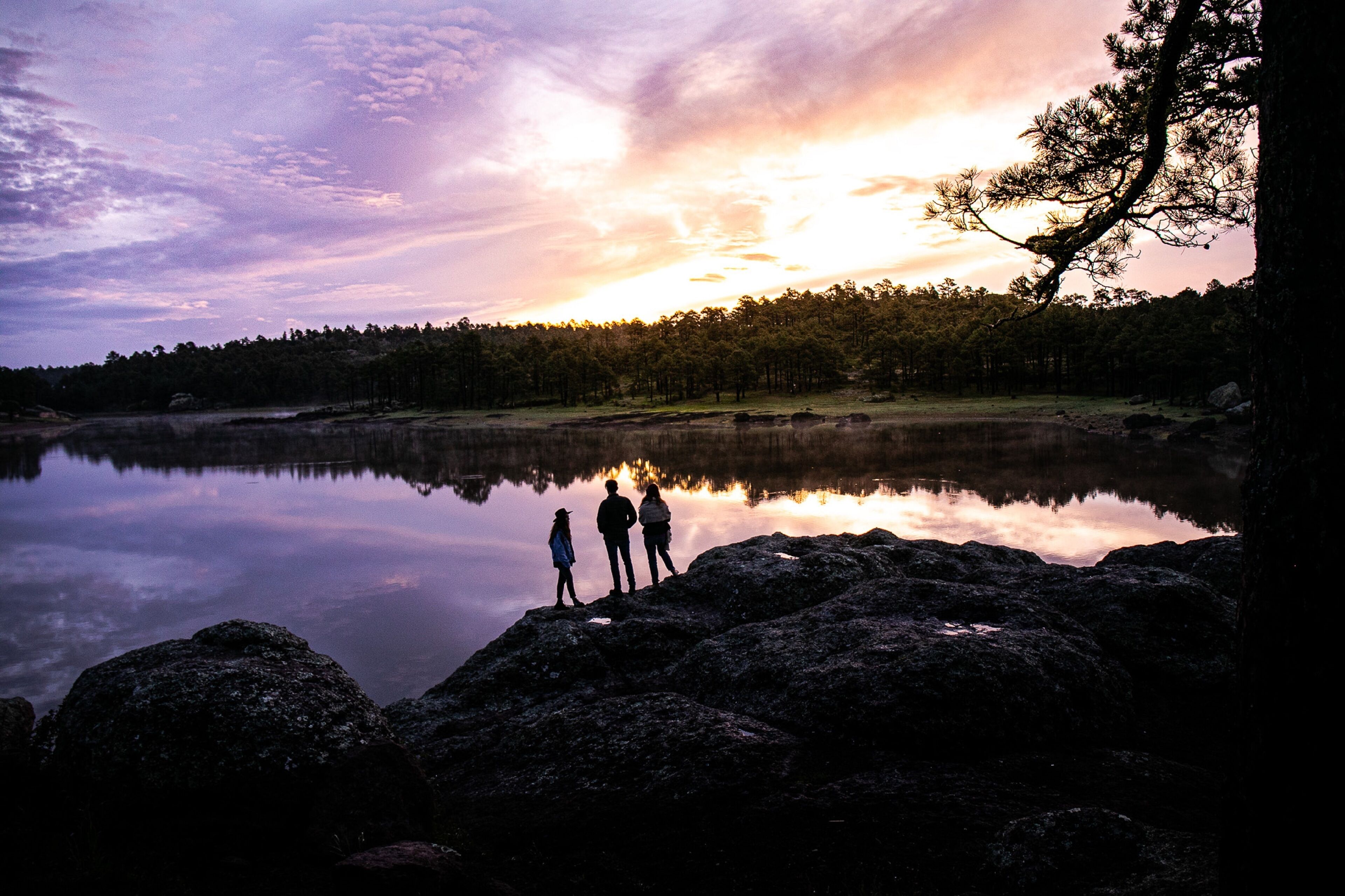 Un paysage paisible au bord d’un lac, avec un coucher de soleil, une eau calme et une forêt en arrière-plan.
