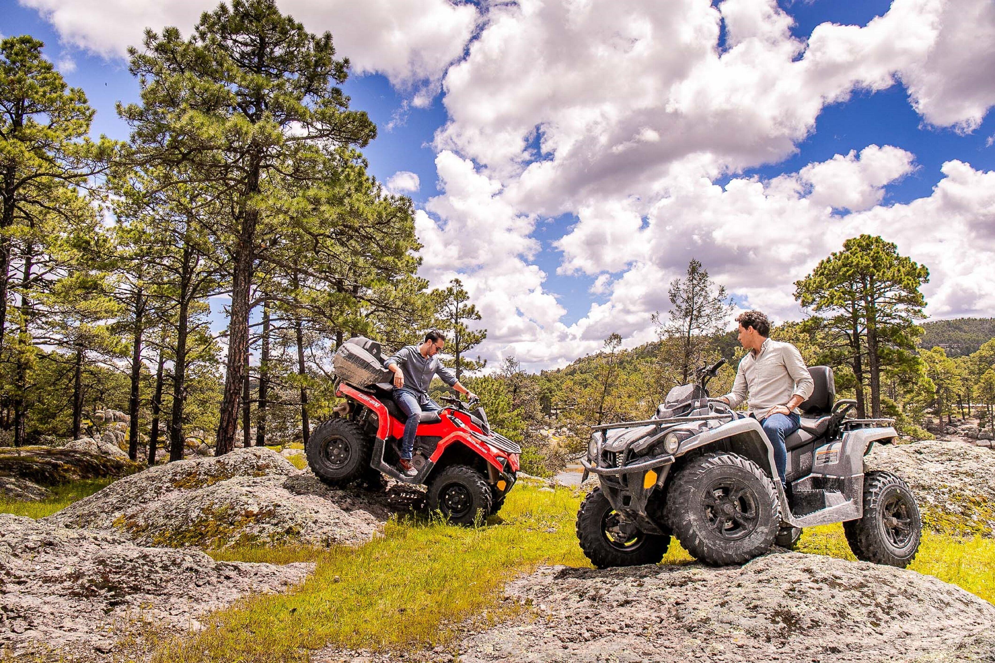 Two people on ATVs in a forested area with rocky terrain and trees.