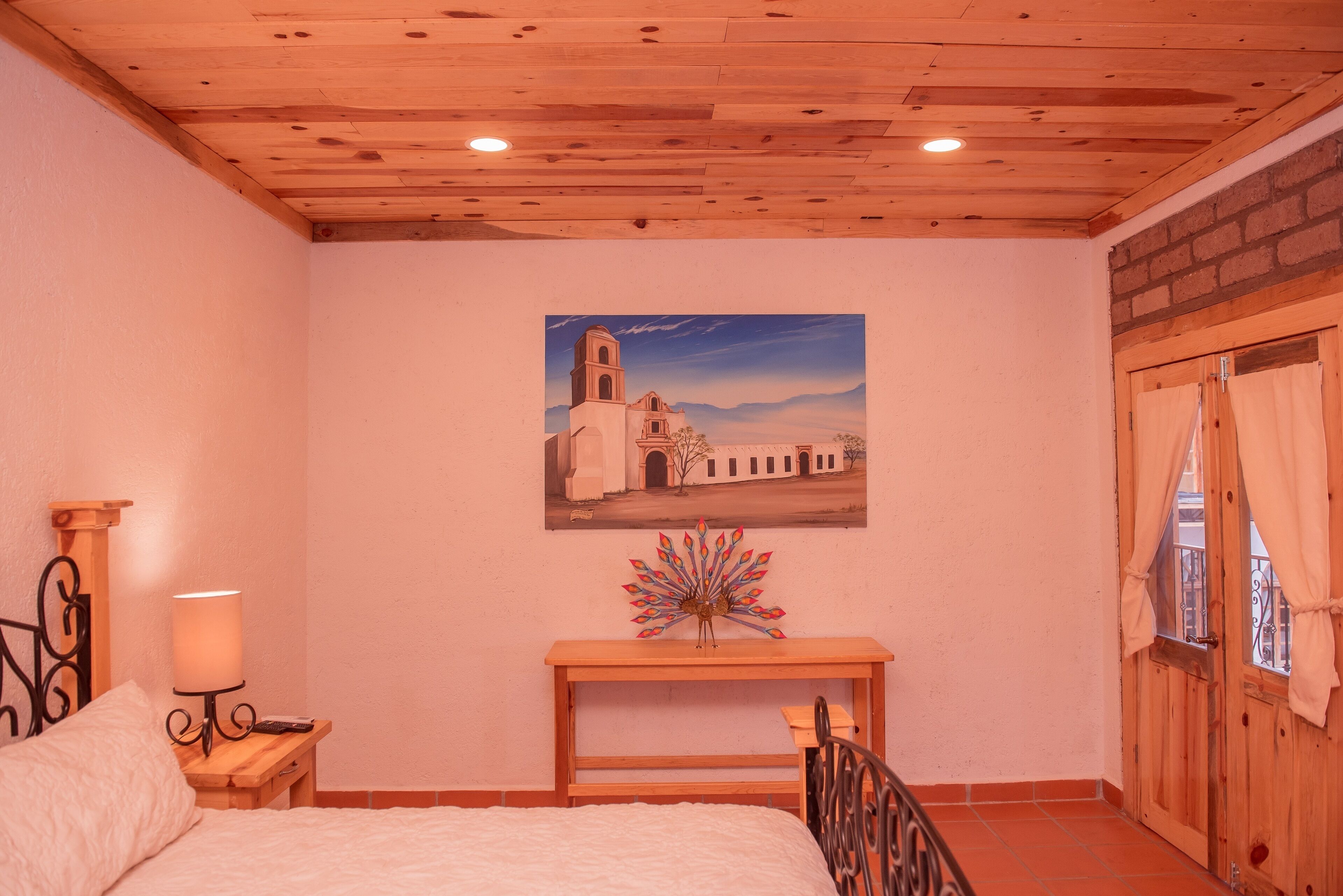 Un dormitorio con techo de madera, una cama con edredón blanco, una mesita de noche con lámpara, una mesa de madera con un objeto decorativo y un cuadro de un edificio en la pared.