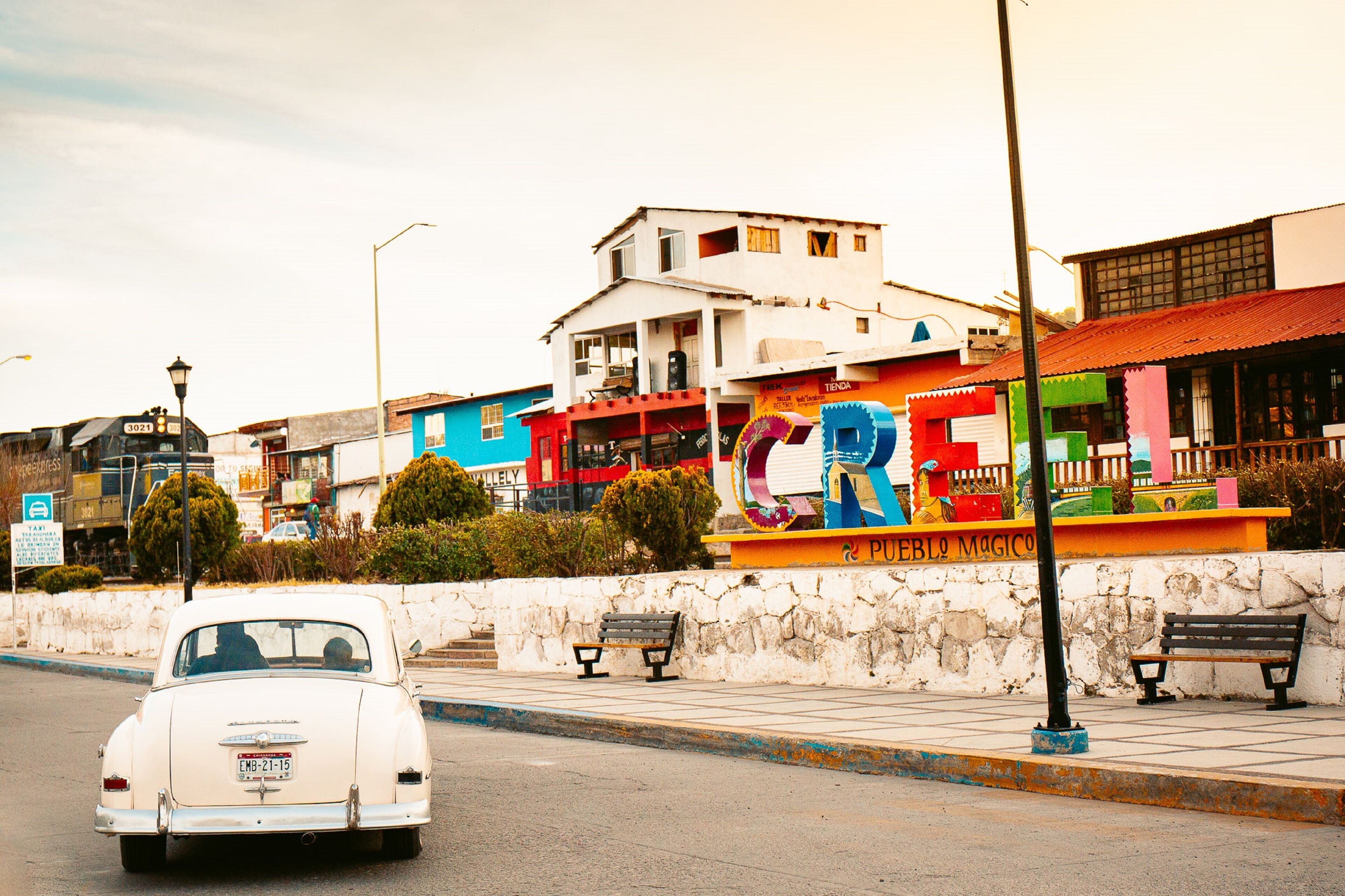 A vintage car driving past a colorful building with a sign that reads "CREO."