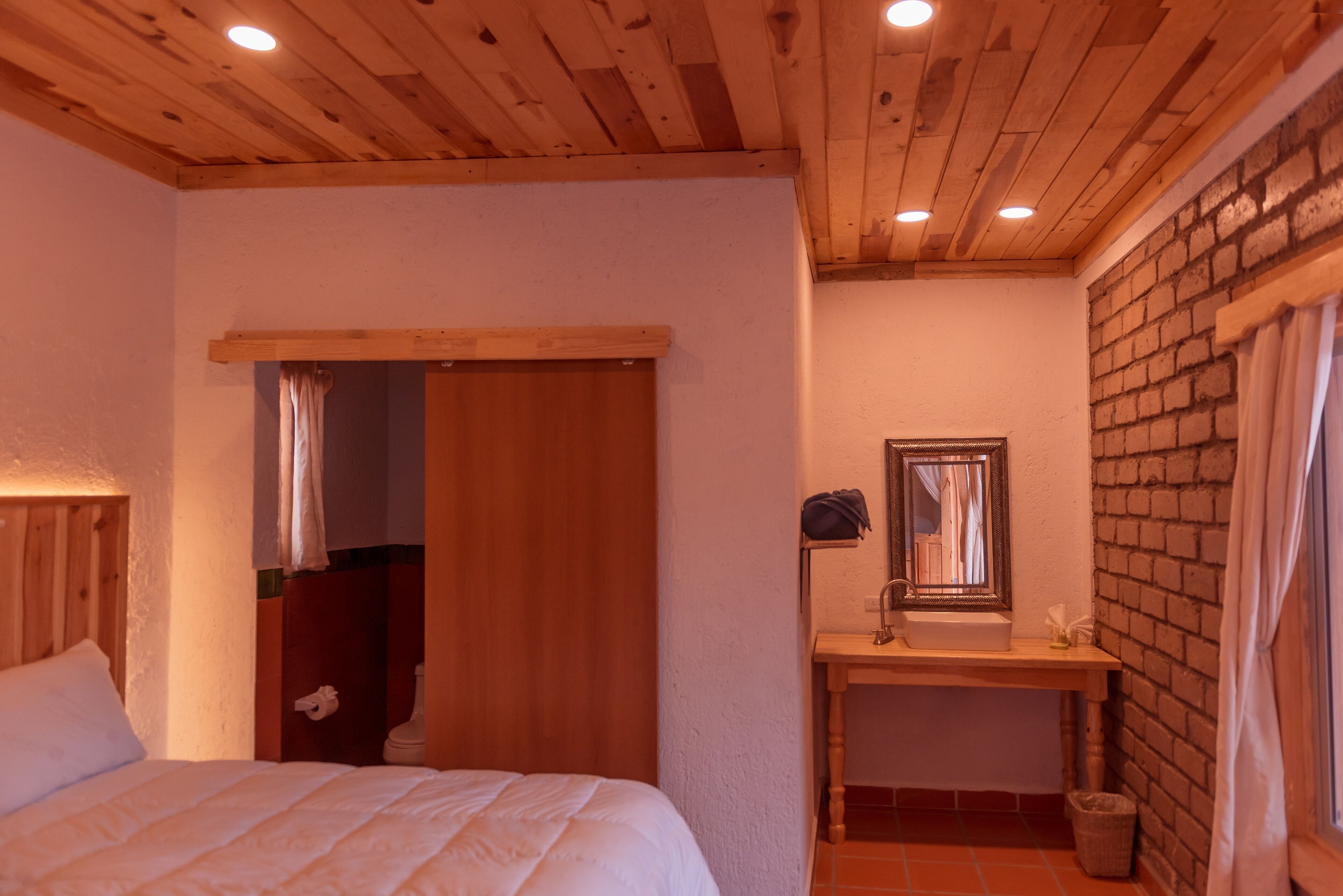 A bedroom with a wooden ceiling, a bed, a wooden door, and a small bathroom with a sink and mirror.