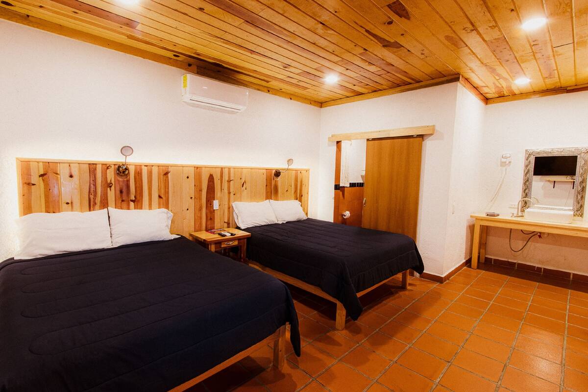 Een kamer met twee bedden, houten hoofdborden, een houten plafond, een betegelde vloer, een klein tafeltje met een spiegel en een deur.