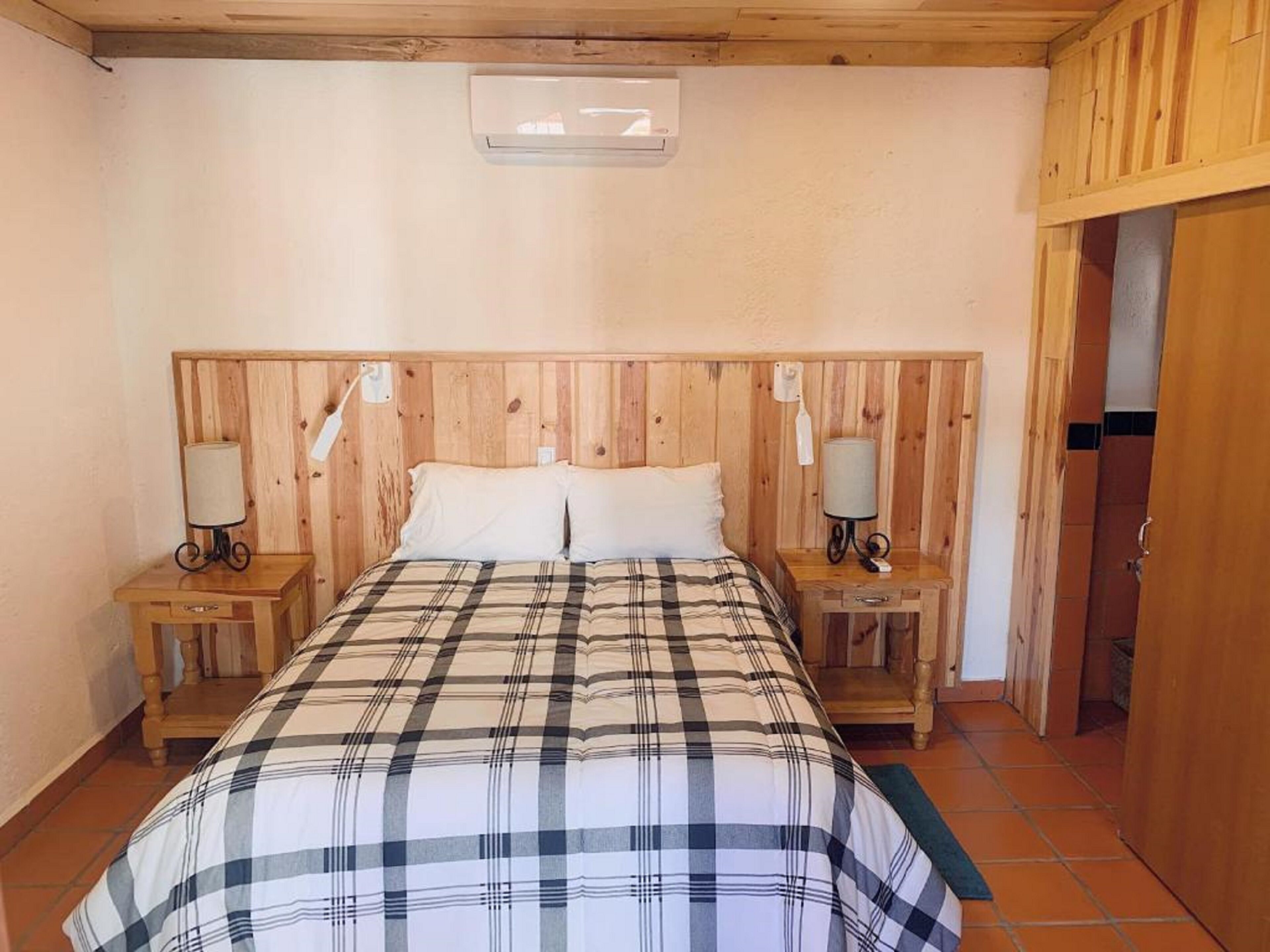 A bedroom with a wooden headboard, two bedside tables with lamps, a plaid bedspread, and a small window above the bed.