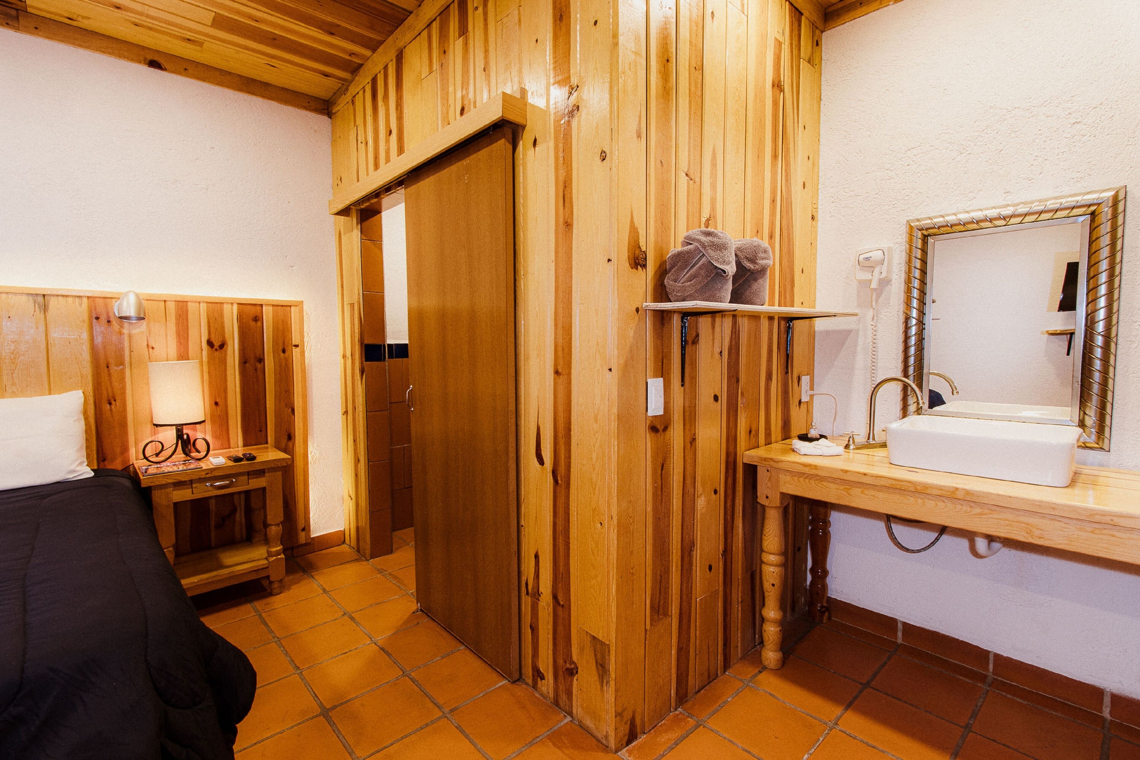 A wooden room with a bed, a nightstand with a lamp, a wooden door, and a bathroom with a sink and mirror.