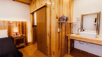 A wooden room with a bed, a nightstand with a lamp, a wooden door, and a bathroom with a sink and mirror.