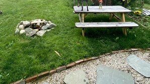 A wooden picnic table with a dog standing nearby, a stone fire pit, and a wooden shed in a grassy backyard. - Cozy cottage loft on the quiet side of Mount Desert Island -Now booking for 2026 (Tremont)