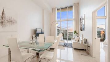 Una sala de estar moderna con una mesa de comedor de vidrio, sofás blancos y un ventanal con vistas a la ciudad.