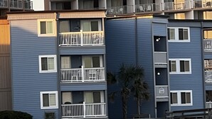A multi-story residential building with balconies, a palm tree, and a sandy ground.