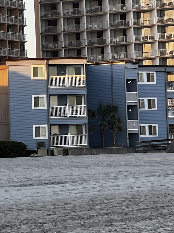 A multi-story residential building with balconies, a palm tree, and a sandy ground.