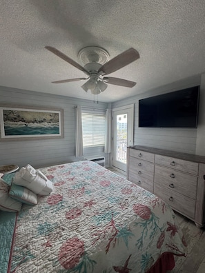 Quarto com cama, ventilador de teto, pintura, janela com persianas, cômoda e televisão fixada na parede.