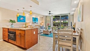 Una cocina moderna con una isla central, armarios de madera y un horno empotrado. Hay una zona de comedor con una mesa y sillas, y una sala de estar con un sofá y una mesita de centro.