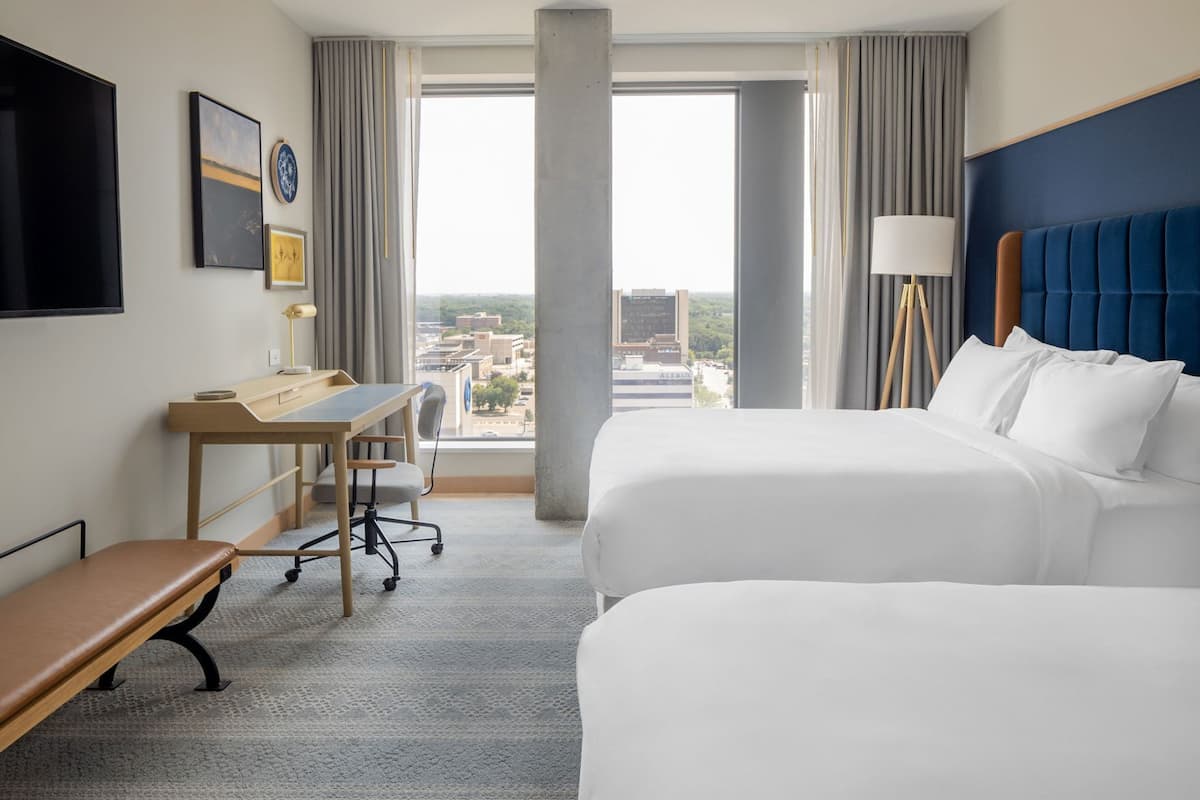A hotel room with two beds, a desk, a chair, a TV, and a view of the city.