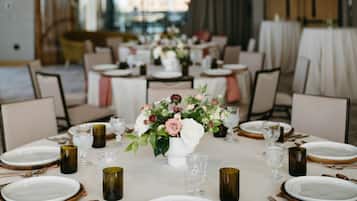Uma sala de jantar com mesas redondas preparadas para uma refeição, com toalhas brancas, pratos com detalhes dourados e um arranjo floral central.