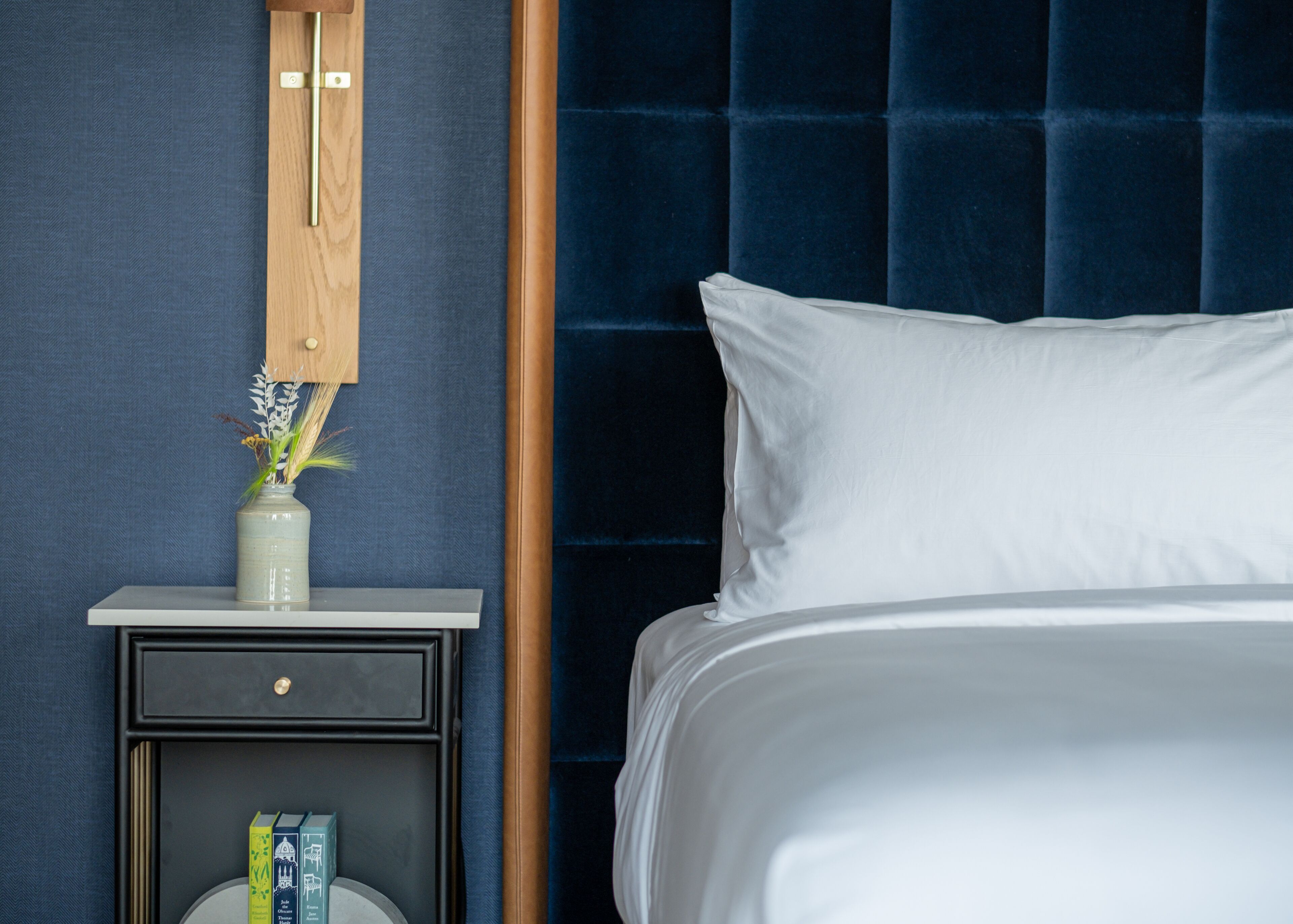 Una cama bien hecha con sábanas blancas, un cabecero azul oscuro y una mesita de noche con un jarrón de flores y un libro.