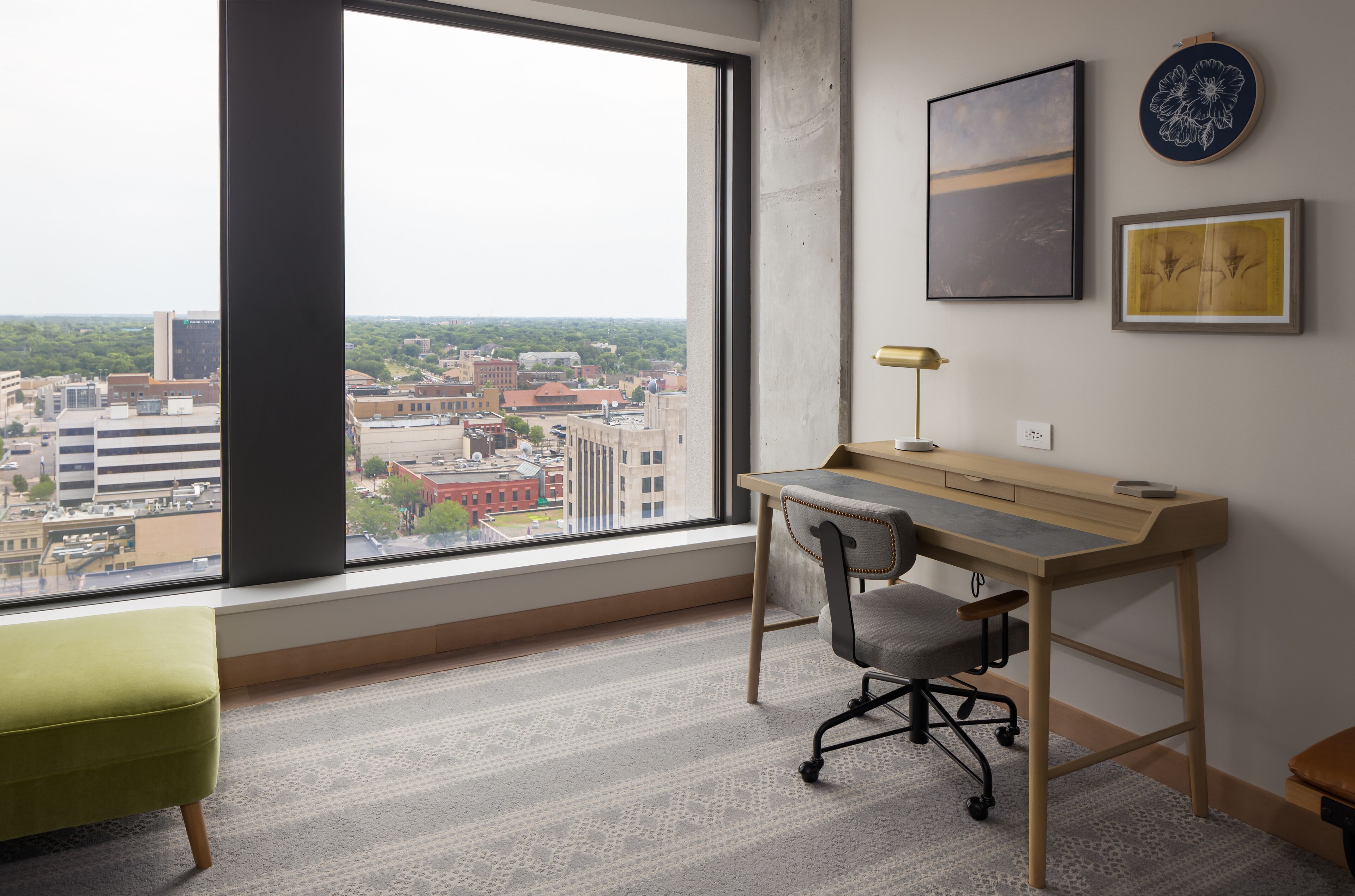 A room with a large window, a wooden desk, a green chair, and a small table.