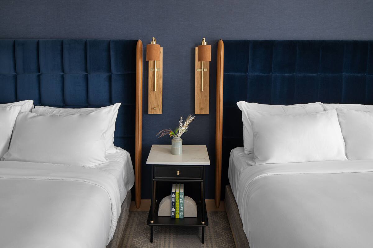 A hotel room with two beds, a dark blue headboard, a small bedside table with a vase, and books.