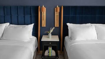 A hotel room with two beds, a dark blue headboard, a small bedside table with a vase, and books.