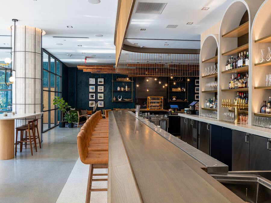 A modern bar area with a long counter, seating, and shelves with bottles and glasses.