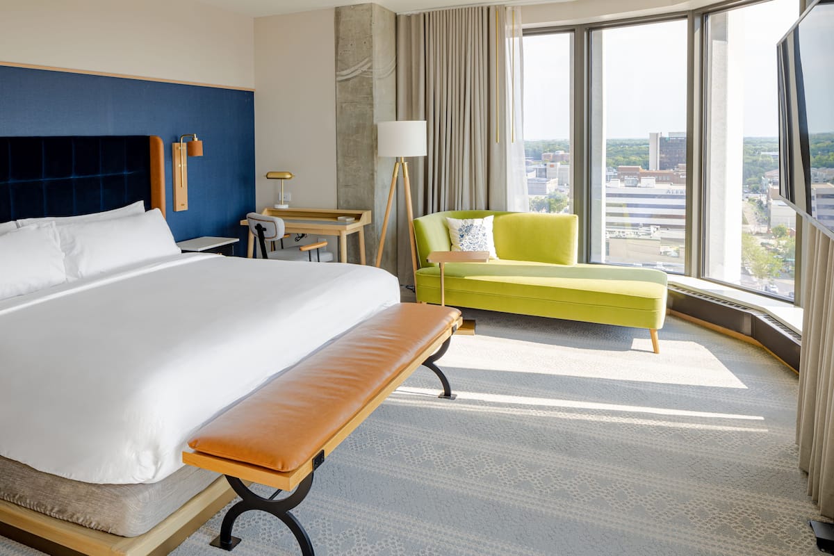 A hotel room with a large bed, a bench, a desk, a chair, and a view of the city through large windows.