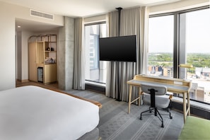 A hotel room with a bed, desk, chair, TV, and a view of the city.