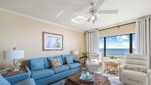 A living room with a blue sofa, a white armchair, a wooden coffee table, and a view of the ocean. - Four Seasons 703E by Liquid Life (Orange Beach)