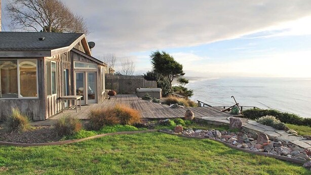A coastal house with a deck overlooking the sea, a stone wall, and a garden area with rocks and plants. - Enter the "Garage House" and step into sublime ocean front living. (Trinidad)
