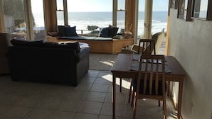A living room with a large bay window overlooking the sea, a sofa, a table, and a wooden deck. - Enter the "Garage House" and step into sublime ocean front living. (Trinidad)
