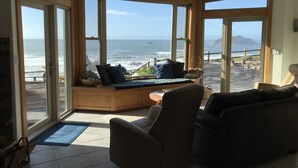 A room with large windows overlooking the ocean, a seating area with cushions, and a ceiling fan. - Enter the "Garage House" and step into sublime ocean front living. (Trinidad)