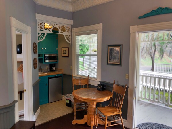 A compact kitchen with teal cabinets, a microwave, and a coffee maker. There's a dining table with chairs and a window with blinds.