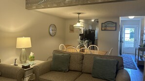 A living room with a couch, a lamp, a clock, a dining table with chairs, and a kitchen in the background. - Sea-Esta Retreat, a Great Golden Isles Location! (St. Simons Island)