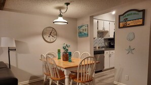 A dining area with a wooden table and white chairs, a clock on the wall, and a kitchen in the background. - Sea-Esta Retreat, a Great Golden Isles Location! (St. Simons Island)