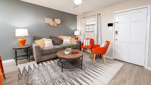 Ein Wohnzimmer mit einem grauen Sofa, zwei orangen Sesseln, einem Couchtisch und einem Deckenventilator.