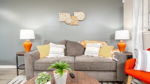 Ein Wohnzimmer mit grauem Sofa, zwei Beistelltischen mit Lampen, einem Couchtisch mit Pflanzen und einem Korb, sowie einem dekorativen Wandbild.