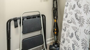 Ein kleines Zimmer mit Deckenventilator, Staubsauger, Klappstuhl aus Metall und einer Wäschetrocknerständer.