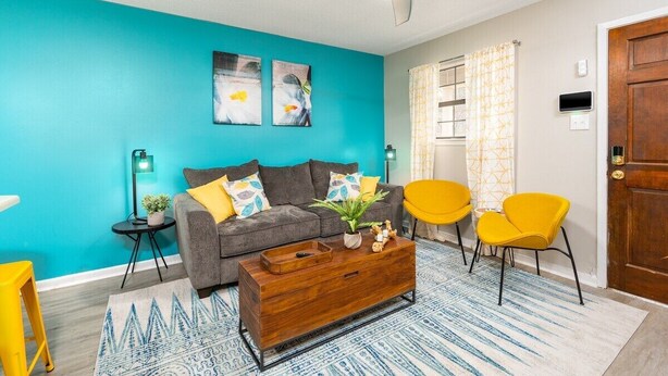 Ein Wohnzimmer mit einer türkisfarbenen Wand, einem grauen Sofa, gelben Sesseln, einem Holztisch und einem Deckenventilator.