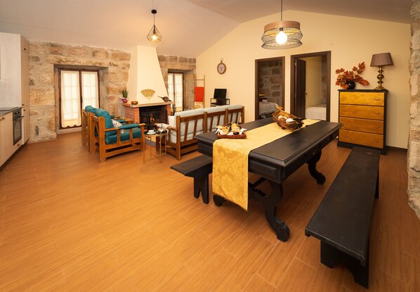 Een gezellige woonkamer met een houten eettafel, bankjes en een bank. Achtergrond is een open haard zichtbaar, en er is een stenen muur.