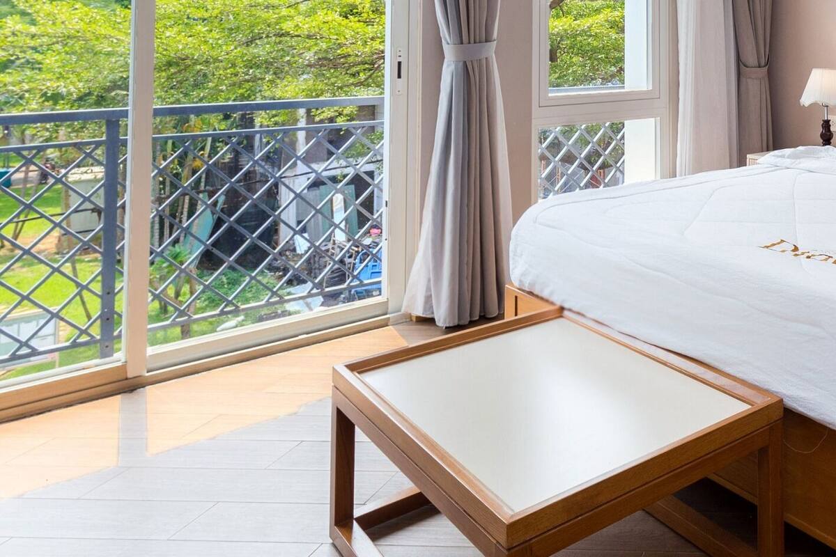 A hotel room with a large bed, a bedside table, a nightstand, and a view of greenery through the window.