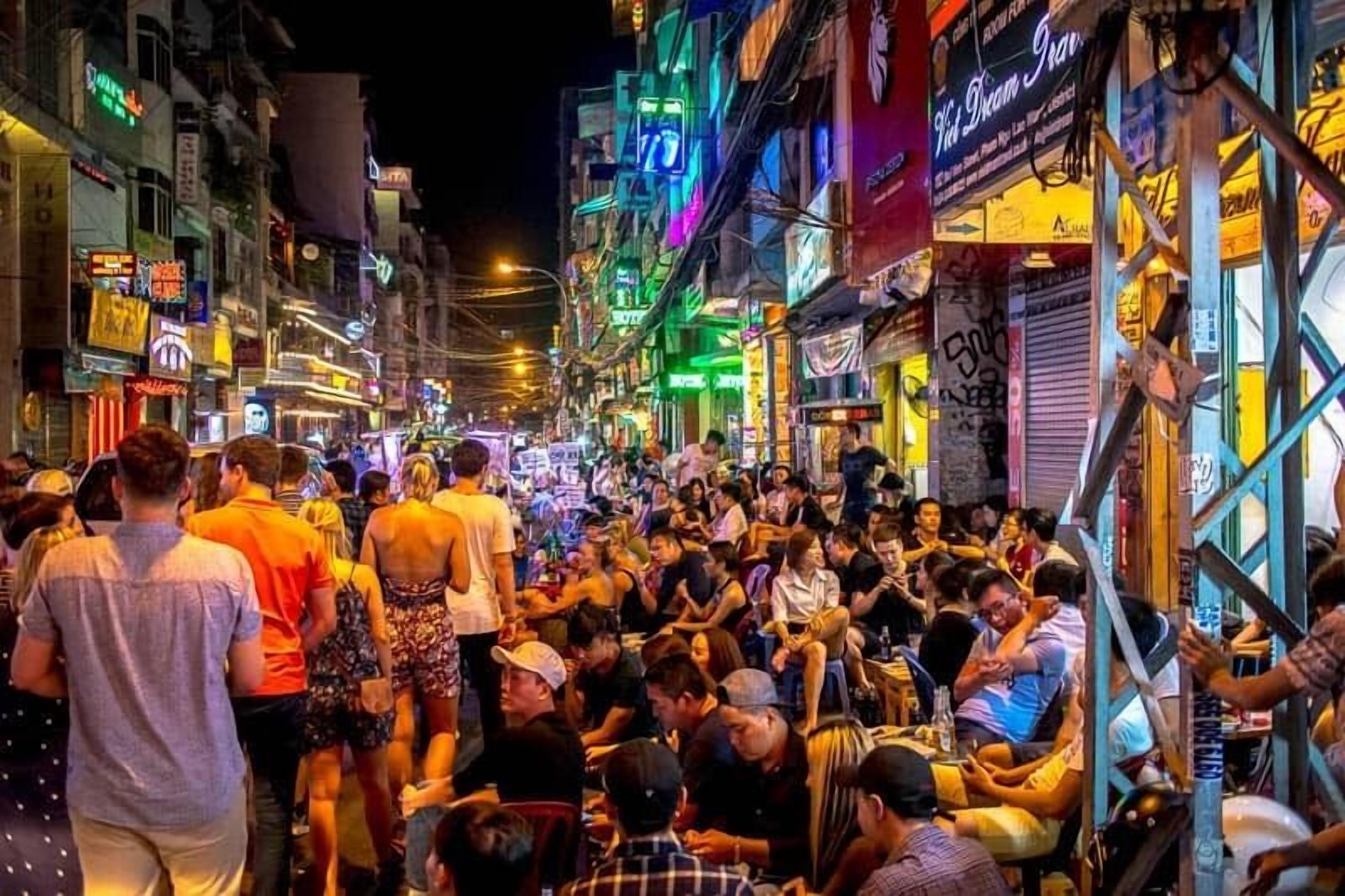 Une rue animée, où l’on voit des gens qui marchent et qui sont assis, entourés de bâtiments illuminés et ornés de diverses enseignes et publicités.