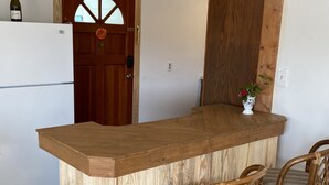 Una cocina con una isla de madera, dos taburetes de mimbre, un refrigerador y una puerta con un ventanal circular.