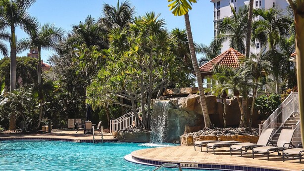 A swimming pool with a waterfall feature, surrounded by palm trees and a stone pathway. - Regatta at Vanderbilt is an upscale condo - a 7 minute walk to the beach! (Naples)