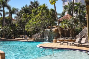 A swimming pool with a waterfall feature, surrounded by palm trees and a stone pathway.