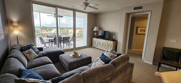 A living room with a gray sectional sofa, a small table, and a TV on a cabinet. There is a balcony with a view of houses and trees.