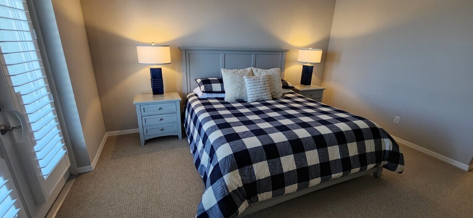 Quarto com colcha xadrez, duas mesas de cabeceira com abajures, um criado-mudo e uma porta com persianas.