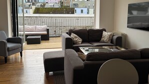 Een moderne woonkamer met een bank, fauteuil en salontafel, een balkon met uitzicht op kleurrijke gebouwen en een aan de muur bevestigde televisie.