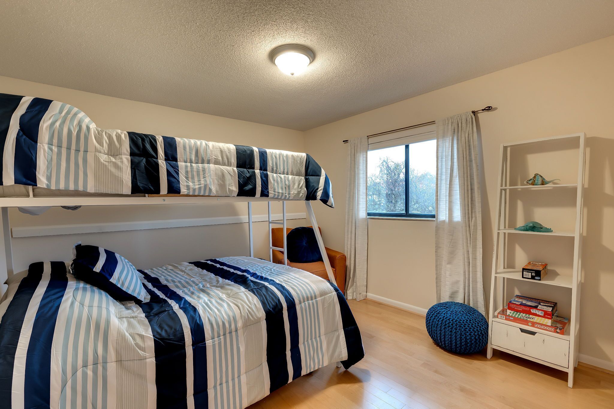 Ein Schlafzimmer mit Etagenbett, einem weißen Kopfteil, einem Fenster mit Vorhängen, einem Bücherregal und einem blauen Sitzsack.