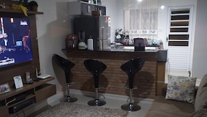 Ein modernes Wohnzimmer mit Fernseher, einer Couch, einem Couchtisch und einer Küchenzeile mit Kühlschrank, Spüle und Barhocker.
