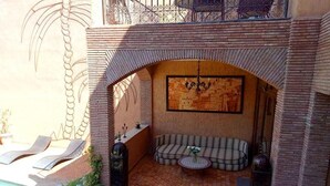 Zona d'estar a l'aire lliure amb sol, una banca amb coixins, una taula rodona petita i una paret decorativa.