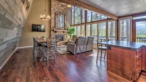 A spacious kitchen with a stone fireplace, wooden ceiling, and large windows offering a view of the outdoors. - Hot Tub, Game Room & Sauna: Ruidoso Hideaway! (Ruidoso)