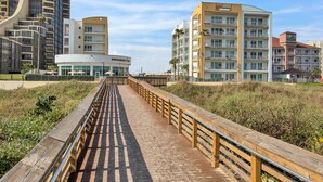 A boardwalk leading to a modern building with a restaurant sign, surrounded by high-rise buildings and palm trees.