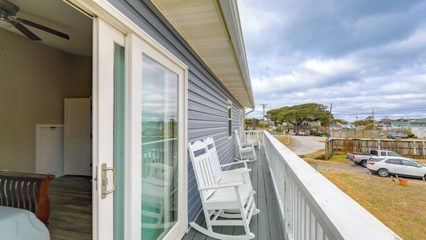 A balcony with white rocking chairs, a sliding glass door, and a view of a residential area with parked cars and houses. - Walkable & Renovated! Salter Path Retreat w/ Deck (Atlantic Beach)